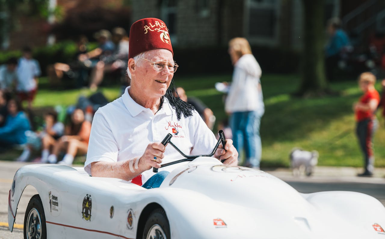 An elderly man in a fez hat drives a toy car during a fun and lively outdoor parade.